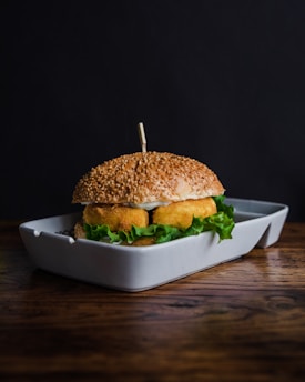 A sesame seed burger bun is filled with crispy, golden-brown fried patties and fresh green lettuce, placed in a white rectangular dish against a dark background. A wooden spike holds the burger together.
