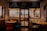 Warmly lit cafe interior with wooden furniture and decorative wall artwork. Three tables are shown, each with different colored chairs. The menu above the door features various items including poultry, salad, and steak. People can be seen sitting inside, engaged in conversation.