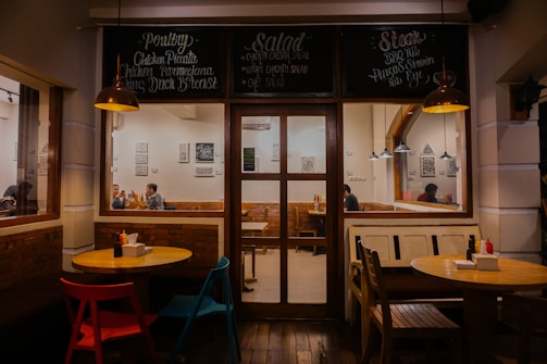 Warmly lit cafe interior with wooden furniture and decorative wall artwork. Three tables are shown, each with different colored chairs. The menu above the door features various items including poultry, salad, and steak. People can be seen sitting inside, engaged in conversation.