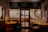 Warmly lit cafe interior with wooden furniture and decorative wall artwork. Three tables are shown, each with different colored chairs. The menu above the door features various items including poultry, salad, and steak. People can be seen sitting inside, engaged in conversation.