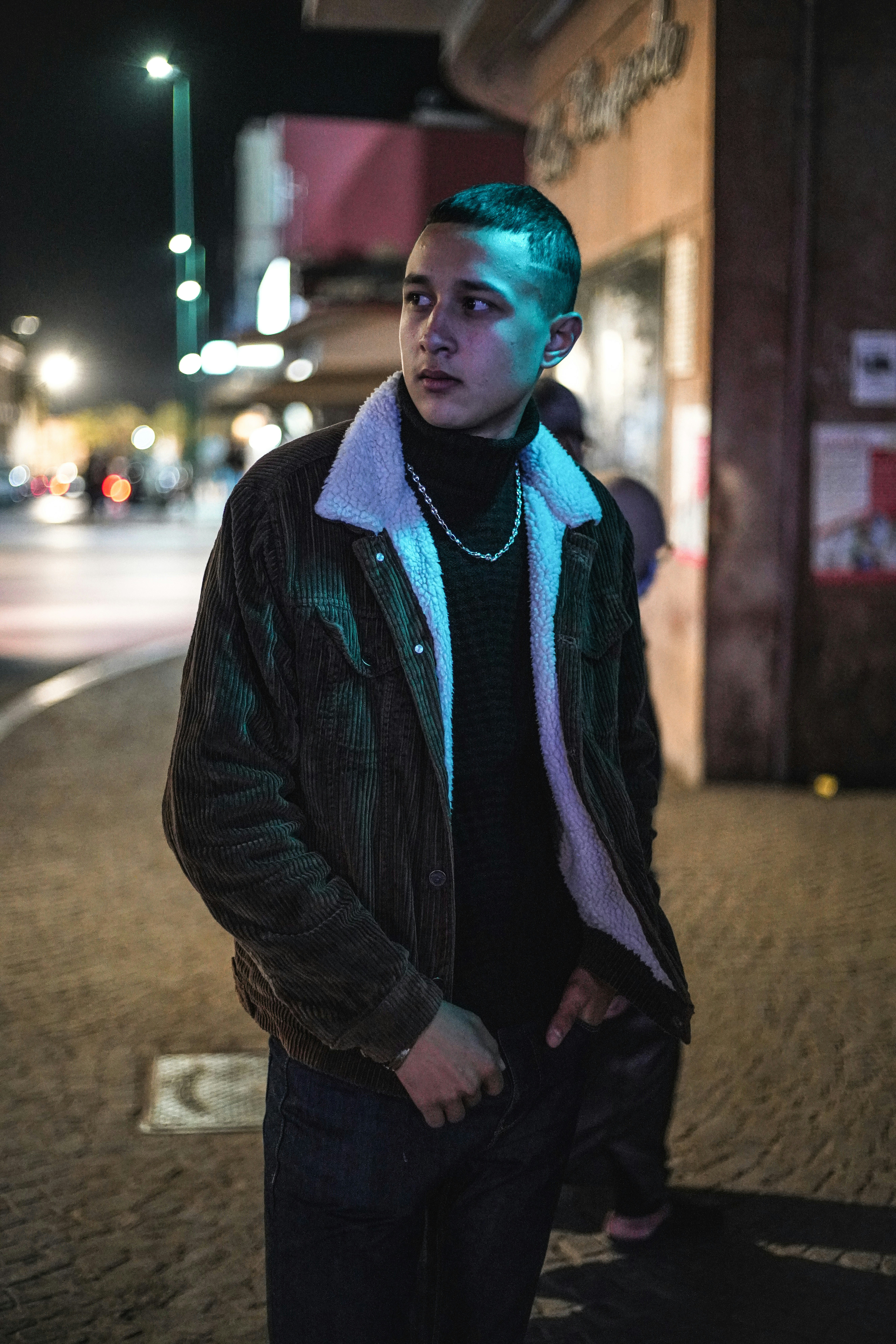 man in black jacket and blue knit cap standing on sidewalk during night time