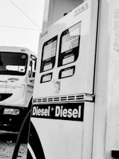 A close-up view of a diesel fuel pump with visible display screens indicating zeros for amounts and a truck parked nearby. The pump has labels displaying 'Diesel' in bold letters.