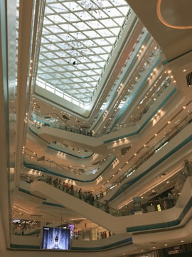 An expansive multi-level shopping mall interior featuring numerous escalators and balconies. The architectural design includes a large glass ceiling that allows natural light to illuminate the space. Several floors are visible with glass railings and retail stores, as well as digital displays and signage indicating different brand stores.