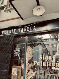 A storefront window displays a sign with 'Klasik Tabela' and advertisements for sign painting and graphic design services. The window reflects nearby buildings and has decorative elements like leaves and a poster with the text 'Consume Obey Die'. The overall setting suggests an artistic or creative atmosphere.