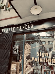 A storefront window displays a sign with 'Klasik Tabela' and advertisements for sign painting and graphic design services. The window reflects nearby buildings and has decorative elements like leaves and a poster with the text 'Consume Obey Die'. The overall setting suggests an artistic or creative atmosphere.