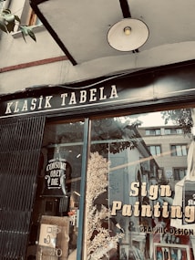 A storefront window displays a sign with 'Klasik Tabela' and advertisements for sign painting and graphic design services. The window reflects nearby buildings and has decorative elements like leaves and a poster with the text 'Consume Obey Die'. The overall setting suggests an artistic or creative atmosphere.