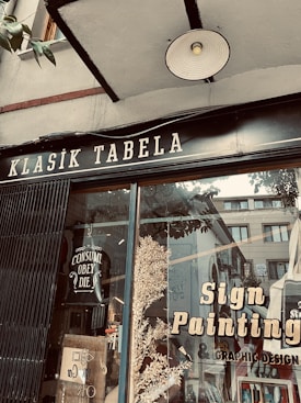 A storefront window displays a sign with 'Klasik Tabela' and advertisements for sign painting and graphic design services. The window reflects nearby buildings and has decorative elements like leaves and a poster with the text 'Consume Obey Die'. The overall setting suggests an artistic or creative atmosphere.
