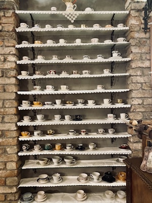 A collection of assorted teacups arranged neatly on multiple shelves against a rustic brick wall. Each shelf is adorned with lace, and the teacups vary in design, color, and size. The arrangement provides a charming and vintage aesthetic.