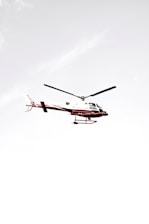 A helicopter flying against a pale, almost white sky with minimal cloud presence. The helicopter has a predominantly white body with red accents and visible rotor blades.