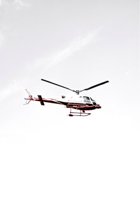 A helicopter flying against a pale, almost white sky with minimal cloud presence. The helicopter has a predominantly white body with red accents and visible rotor blades.