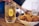 A chilled glass of Suntory Whisky filled with ice sits on a wooden table next to a bowl of crispy, thinly sliced lotus root chips. Chopsticks are picking up one of the chips, emphasizing a relaxed, casual dining atmosphere.