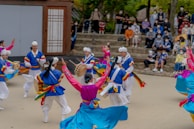 A group of performers dressed in traditional Korean attire are engaged in a lively, colorful dance in front of an outdoor audience. The dancers wear vibrant costumes with hues of blue, pink, yellow, and white, and some are playing traditional percussion instruments. The audience, seated on stone steps, observes the performance attentively.