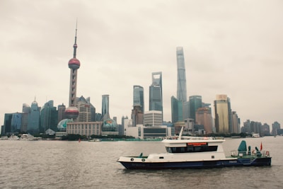 A large city skyline with modern skyscrapers and the iconic Oriental Pearl Tower dominating the view. The buildings are set against a cloudy sky. In the foreground, a police boat is seen on the river, adding a dynamic element to the urban landscape.