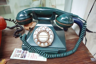 A vintage rotary dial telephone with a dark green body and handset. The phone has a coiled cord and is set on a wooden surface next to an orange telephone. There is a piece of paper with printed text and images placed near the phones.
