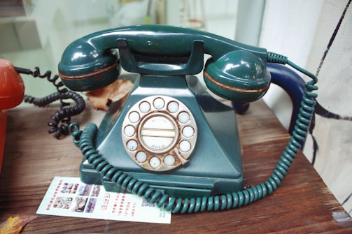 A vintage rotary dial telephone with a dark green body and handset. The phone has a coiled cord and is set on a wooden surface next to an orange telephone. There is a piece of paper with printed text and images placed near the phones.