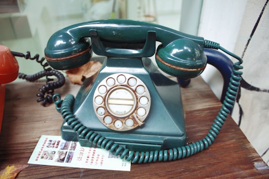 A vintage rotary dial telephone with a dark green body and handset. The phone has a coiled cord and is set on a wooden surface next to an orange telephone. There is a piece of paper with printed text and images placed near the phones.