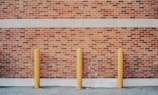 Three yellow bollards are evenly spaced in front of a red brick wall, which includes a cream-colored stripe near the top. The ground is covered with gravel, and there is a visible shadow cast on the wall.