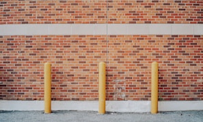 Three yellow bollards are evenly spaced in front of a red brick wall, which includes a cream-colored stripe near the top. The ground is covered with gravel, and there is a visible shadow cast on the wall.
