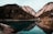 A serene mountain landscape with a clear lake reflecting the towering cliffs and dense pine forests. A small bridge and a house are nestled on the right side near the water's edge.