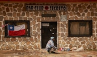 A person, dressed in a blue and white striped sweater and blue beanie, is seen squatting in front of a stone-walled building with a sign that reads 'BAR CLUB SECRETO'. A Chilean flag is displayed in the window to the left of the door, and several bags are placed on the ground near the person. The building has barred windows and the door has a sign with a phone number and address on it.