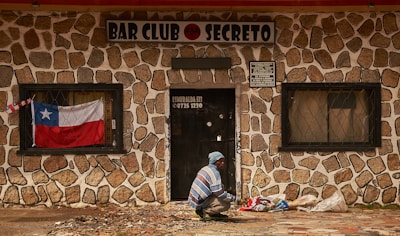 A person, dressed in a blue and white striped sweater and blue beanie, is seen squatting in front of a stone-walled building with a sign that reads 'BAR CLUB SECRETO'. A Chilean flag is displayed in the window to the left of the door, and several bags are placed on the ground near the person. The building has barred windows and the door has a sign with a phone number and address on it.