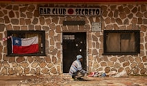 A person, dressed in a blue and white striped sweater and blue beanie, is seen squatting in front of a stone-walled building with a sign that reads 'BAR CLUB SECRETO'. A Chilean flag is displayed in the window to the left of the door, and several bags are placed on the ground near the person. The building has barred windows and the door has a sign with a phone number and address on it.