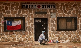 A person, dressed in a blue and white striped sweater and blue beanie, is seen squatting in front of a stone-walled building with a sign that reads 'BAR CLUB SECRETO'. A Chilean flag is displayed in the window to the left of the door, and several bags are placed on the ground near the person. The building has barred windows and the door has a sign with a phone number and address on it.