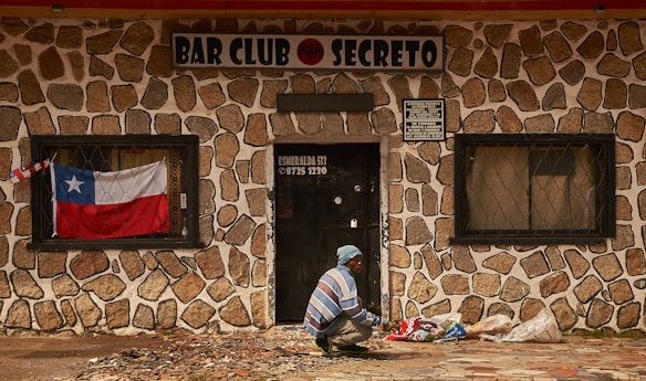 A person, dressed in a blue and white striped sweater and blue beanie, is seen squatting in front of a stone-walled building with a sign that reads 'BAR CLUB SECRETO'. A Chilean flag is displayed in the window to the left of the door, and several bags are placed on the ground near the person. The building has barred windows and the door has a sign with a phone number and address on it.