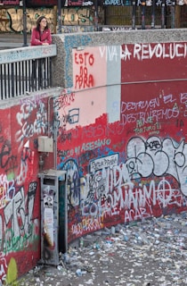 A person stands on a ledge overlooking a heavily graffitied wall covered in a multitude of colorful messages and tags. The ground below is littered with trash and debris, suggesting an urban setting with signs of unrest or protest. The graffiti contains political messages in various languages.