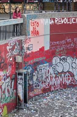 A person stands on a ledge overlooking a heavily graffitied wall covered in a multitude of colorful messages and tags. The ground below is littered with trash and debris, suggesting an urban setting with signs of unrest or protest. The graffiti contains political messages in various languages.