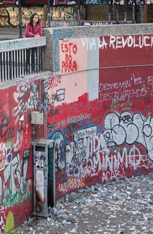 A person stands on a ledge overlooking a heavily graffitied wall covered in a multitude of colorful messages and tags. The ground below is littered with trash and debris, suggesting an urban setting with signs of unrest or protest. The graffiti contains political messages in various languages.