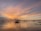 Traditional fishing nets extend over a calm body of water at sunset, with a sky painted in hues of orange, pink, and purple. The structures supporting the nets are silhouetted against the vibrant sky, reflecting on the water's surface.