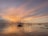 Traditional fishing nets extend over a calm body of water at sunset, with a sky painted in hues of orange, pink, and purple. The structures supporting the nets are silhouetted against the vibrant sky, reflecting on the water's surface.
