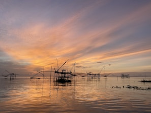 Traditional fishing nets extend over a calm body of water at sunset, with a sky painted in hues of orange, pink, and purple. The structures supporting the nets are silhouetted against the vibrant sky, reflecting on the water's surface.