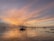Traditional fishing nets extend over a calm body of water at sunset, with a sky painted in hues of orange, pink, and purple. The structures supporting the nets are silhouetted against the vibrant sky, reflecting on the water's surface.