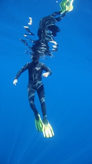 A person is underwater wearing a black wetsuit and bright yellow fins. The reflection of the swimmer is visible on the water surface above, with light playing off the vibrant blue surroundings.