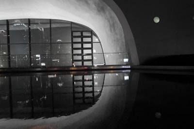A modern architectural structure featuring a large window with grid patterns reflects on a still body of water. The soft glow of city lights is visible through the glass, creating a serene ambiance. The interior has smooth, curved walls, and the overall atmosphere is dimly lit, suggesting nighttime.