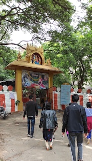 A group of people walking towards an entrance adorned with an intricate golden arch featuring statues of deities. The entrance is decorated with a painted depiction of a religious scene above a red and white striped wall. Lush green trees surround the area, creating a serene atmosphere.