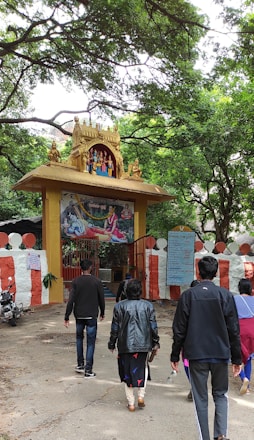 A group of people walking towards an entrance adorned with an intricate golden arch featuring statues of deities. The entrance is decorated with a painted depiction of a religious scene above a red and white striped wall. Lush green trees surround the area, creating a serene atmosphere.