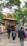 A group of people walking towards an entrance adorned with an intricate golden arch featuring statues of deities. The entrance is decorated with a painted depiction of a religious scene above a red and white striped wall. Lush green trees surround the area, creating a serene atmosphere.