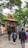 A group of people walking towards an entrance adorned with an intricate golden arch featuring statues of deities. The entrance is decorated with a painted depiction of a religious scene above a red and white striped wall. Lush green trees surround the area, creating a serene atmosphere.