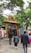 A group of people walking towards an entrance adorned with an intricate golden arch featuring statues of deities. The entrance is decorated with a painted depiction of a religious scene above a red and white striped wall. Lush green trees surround the area, creating a serene atmosphere.