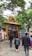A group of people walking towards an entrance adorned with an intricate golden arch featuring statues of deities. The entrance is decorated with a painted depiction of a religious scene above a red and white striped wall. Lush green trees surround the area, creating a serene atmosphere.