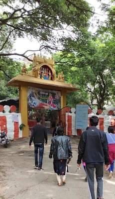 A group of people walking towards an entrance adorned with an intricate golden arch featuring statues of deities. The entrance is decorated with a painted depiction of a religious scene above a red and white striped wall. Lush green trees surround the area, creating a serene atmosphere.