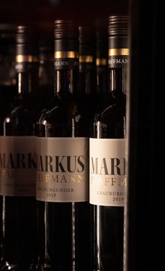 Several bottles of wine with the labels prominently displaying the name 'Markus Pfaffmann' and 'Grauburgunder 2019', arranged in a dimly lit environment.