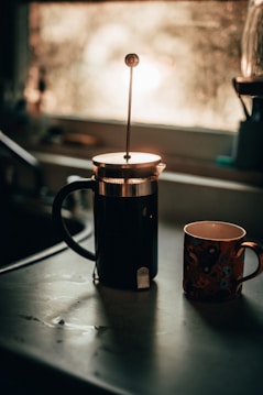 A French press coffee maker and a decorative ceramic mug stand on a kitchen counter. The scene is bathed in soft, warm light from a nearby window, giving a cozy and inviting atmosphere. In the background, elements of the kitchen including the sink and faucet are visible but out of focus.