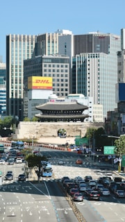 A busy urban scene featuring a historic gate surrounded by modern high-rise buildings. The street is filled with vehicles, including cars and buses, and the area is bustling with traffic. Billboards and advertisements are visible on the buildings, and the scene is bright with clear skies.