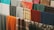 A collection of various colorful fabrics and textiles are hung in a row. The fabrics display a range of patterns, including plaid, floral, and solid designs. Materials appear to be made of different textures, such as wool or cotton, in an array of colors like orange, blue, green, and red.
