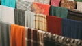 A collection of various colorful fabrics and textiles are hung in a row. The fabrics display a range of patterns, including plaid, floral, and solid designs. Materials appear to be made of different textures, such as wool or cotton, in an array of colors like orange, blue, green, and red.