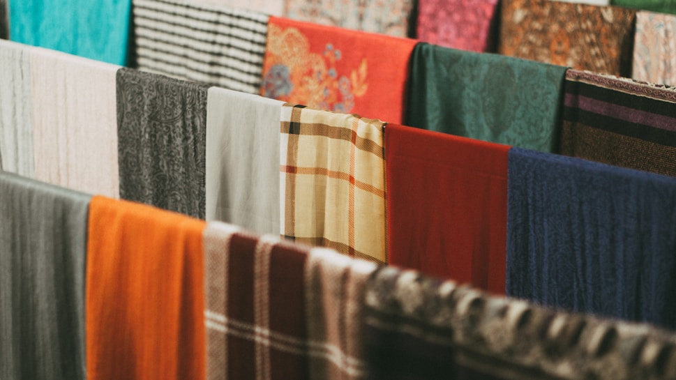 A collection of various colorful fabrics and textiles are hung in a row. The fabrics display a range of patterns, including plaid, floral, and solid designs. Materials appear to be made of different textures, such as wool or cotton, in an array of colors like orange, blue, green, and red.